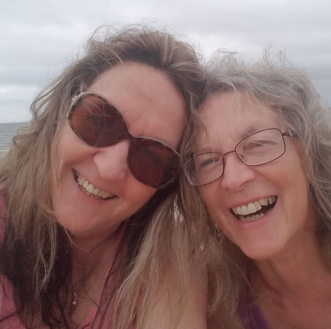 Close-up selfie of two smiling women on a grey-skied beach day